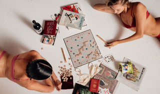 Two women wearing lingerie playing a board game on a white floor with various items around them.
