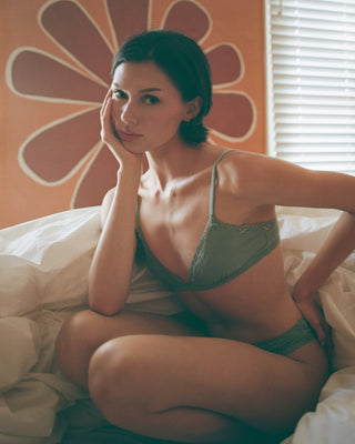 Woman in a green bra and panties sitting on a bed with a floral wall in the background