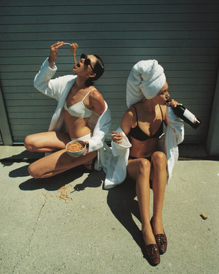 Two women in bathrobes and underwear sitting on a concrete surface, one eating and the other drinking.