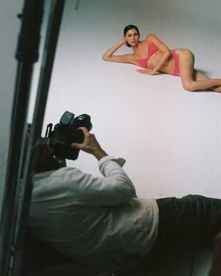 Model in pink Love and War microfiber bralette and panties lying on a white floor with a photographer taking pictures.